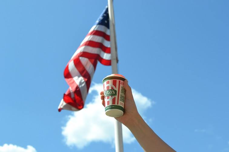 A hand holds up a Rita’s branded cup against an American flag against a blue sky with a couple of fluffy white clouds.