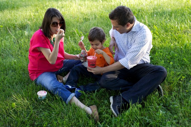 A pregnant woman and her husband sit cross-legged on the grass with their toddler son between them, all sharing some frozen Rita’s treats.