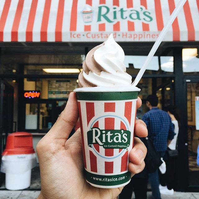 A hand holds a Rita’s branded cup with frozen custard swirl on top in front of a Rita’s store.