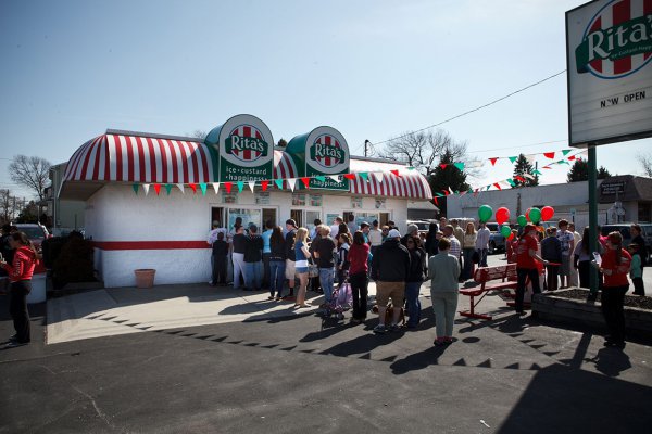 Double lines of people are queued up at a Rita’s. Red, white and green bunting hangs over the parking lot, and red and green balloons are seen in the background.