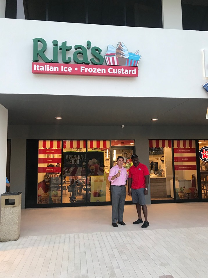 Two men stand in front of a Rita’s shop in a strip mall. The man on the left is holding a chocolate cone. A sign above the shop reads “Rita’s” and “Italian Ice” and “Frozen Custard.”