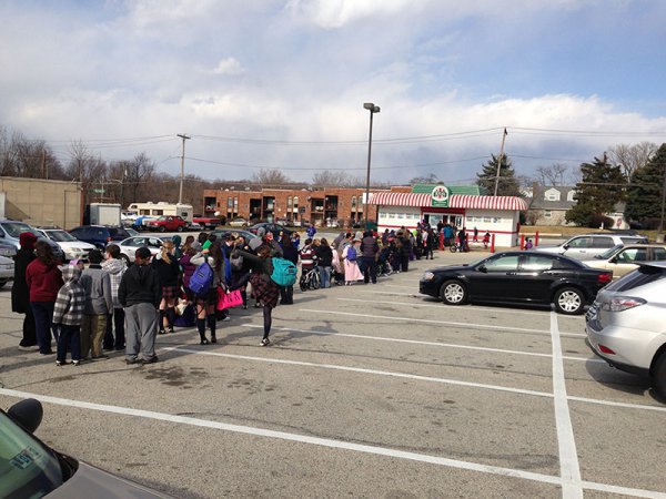 Two lines of people in winter coats stretch across a parking lot in front of a Rita's Italian Ice franchise.
