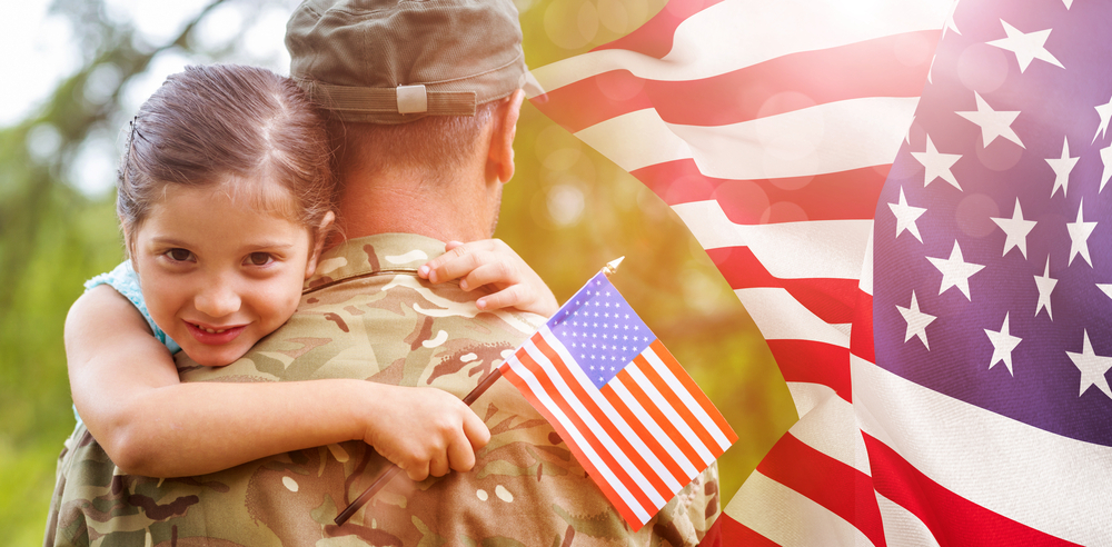 A little girl clutches a small American flag while she hugs her father, who is wearing combat fatigues. A large American flag waves in the background on the right-hand side of the photo.